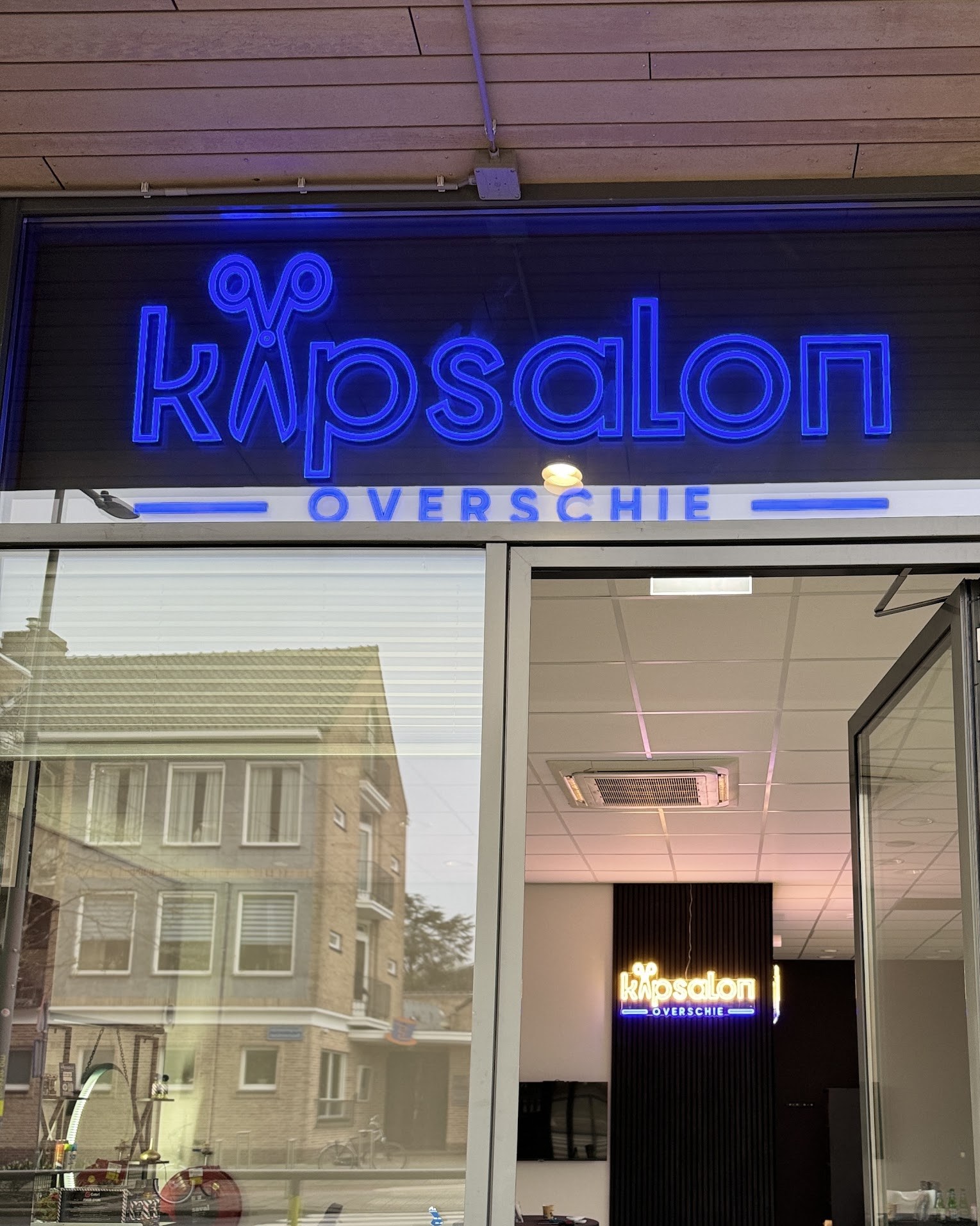De gevel van Kapsalon Overschie met het blauwe neonbord boven de open deur